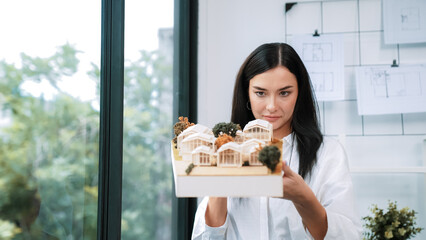 Close up of skilled young beautiful caucasian architect lift house model left side and right sight to check and focus on inspecting mistake point at modern office with blueprint. Immaculate.
