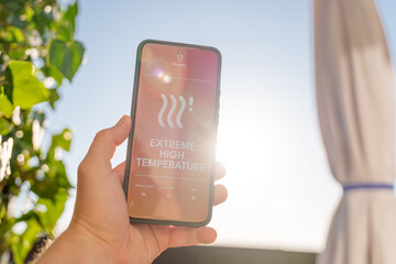 Hand holding smartphone showing extreme high temperature alert on weather app with bright sunny sky...