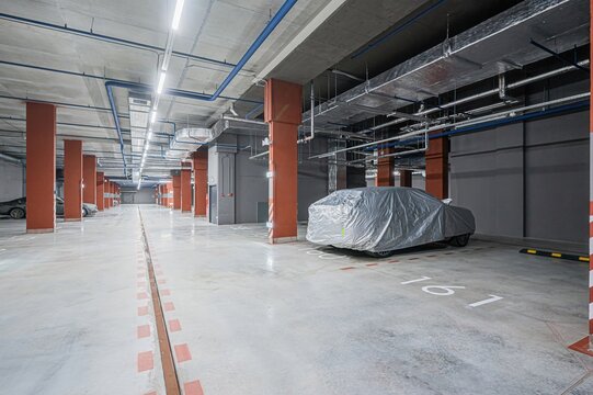 underground parking garage. A car covered in silver sits in its spot.  Pipes line the ceiling above rows of reddish-brown columns