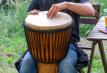 man playing a traditional djembe drum outdoors, sitting next to a wooden table in a natural green...