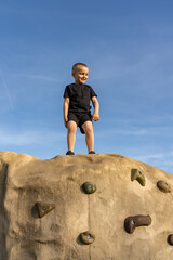 Little boy standing on top of a rock climbing mountain against a blue sky background. Sports and...