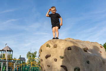 Happy little boy stands on top of an artificial climbing mountain he climbed against a blue sky....