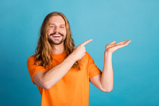 Confident young man with long hair wearing orange t-shirt presenting with hand gesture against blue background - Powered by Adobe