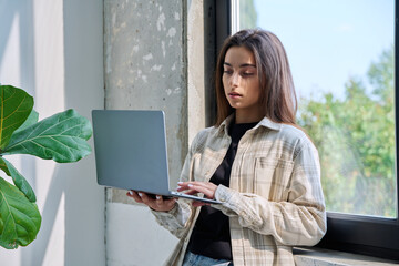 Portrait of young woman standing with laptop computer in home interior near window