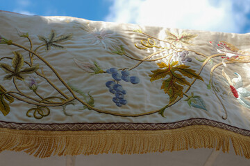 Close-up of an ornate embroidered processional canopy used in Catholic religious ceremonies, particularly during the Feast of Corpus Christi. Blue sky enhances the sacred feel. © Ganna Zelinska