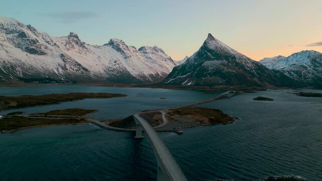 Ramberg auf den Lofoten in Norwegen bei Sonnenuntergang im Winter, Stra&szlig;e f&uuml;hrt &uuml;ber Br&uuml;cke zum schneebedeckten Berg, weiches Licht &uuml;ber K&uuml;ste, Meer und felsiger arktischer Landschaft