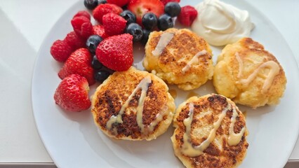 Golden cheese pancakes served with fresh strawberries, raspberries, blueberries and a dollop of cream on a white plate. The image represents a sweet breakfast, indulgence and homemade comfort
