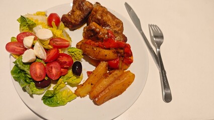 JJuicy roasted ribs with baked potatoes and peppers served alongside a fresh salad with tomatoes, mozzarella and olives. The image represents hearty homemade cooking and balanced dining