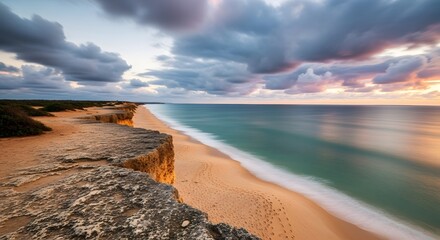 Dramatic coastal view with golden sand and turquoise water under a cloudy sky during sunset. Cliffside vegetation contrasts with the beach. Soft sunlight creates a tranquil atmosphere.