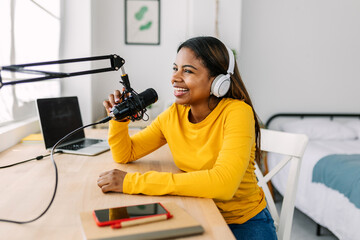 Happy young woman speaking into a microphone, recording her podcast from the comfort of her home,...