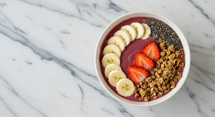 A&ccedil;a&iacute; bowl with sliced bananas, strawberries, granola, and chia seeds arranged in a white bowl on a marble surface.