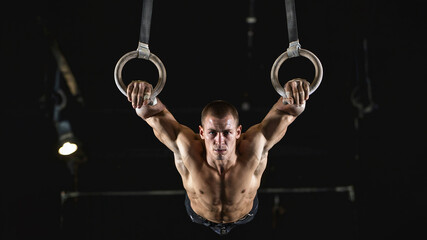Male gymnast performing a strength move on gymnastic rings in a dark gym. Intense expression, muscular body, dramatic spotlighting. Front angle with black background.