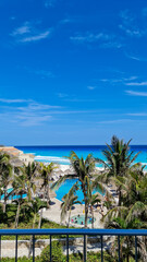 A stunning tropical view of a seaside paradise, featuring lush palm trees, a light blue pool, and crystalline ocean waves under a sunny sky. The perfect beach day.