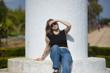Young woman, beautiful, blonde, wearing a black t-shirt, jeans, and sunglasses, sitting on white...