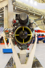 Helicopter gas turbine engine on display at aviation exhibition. Engine is mounted on stand, showcasing its intricate mechanical components and turbine blades. Colorful helicopter in background