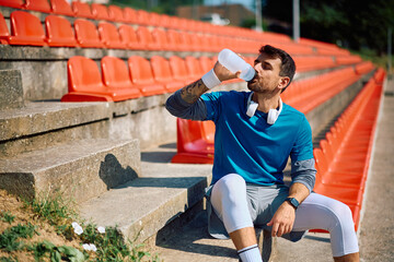 Male athlete drinking water while relaxing on staircase at stadium.