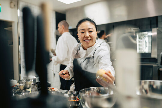 Smiling female head chef reaching for grabbing vegetable while working in restaurant kitchen