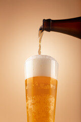 Golden beer pours from bottle into tall glass, creating thick foam and visible bubbles. Warm...