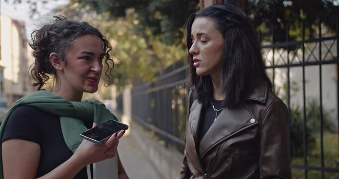 Two young female friends engage in a serious and candid conversation outdoors. One friend is seen holding a smart phone, possibly recording audio or receiving an important call.