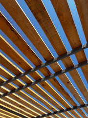 Modern wooden pergola roof with evenly spaced slats against a clear blue sky. The architectural structure creates a dynamic geometric pattern with light and shadow, highlighting natural wood texture