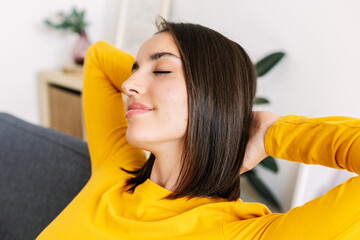 Closeup portrait of young beautiful woman lying on couch at home. Millennial pretty female with hands over head relaxing on sofa- Well-being concept.