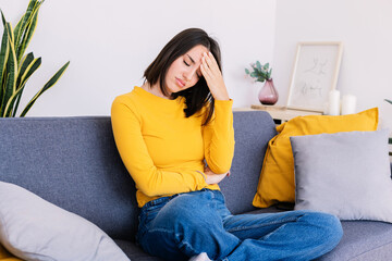 Young woman sitting on sofa at home suffering a headache. Adult female touching forehead having a...