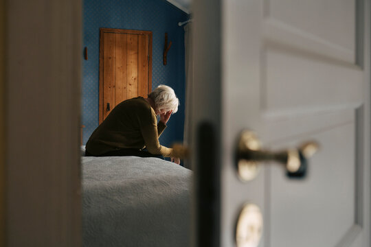 Depressed senior woman sitting with face down on bed seen through bedroom door