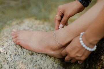 Woman fastens a heart-shaped anklet on her ankle while sitting by a river, showing off a pearl...