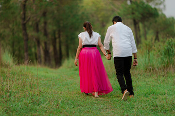 Naklejka premium Couple strolling hand-in-hand through a grassy meadow, enjoying nature. She wears a vibrant pink skirt, adding a pop of color. Back view.
