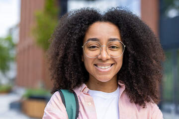 A smiling young woman with curly hair wearing glasses and a pink shirt. She has a backpack,...