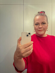 Midlife woman with gray hair wearing a red shirt, taking a selfie in a bright bathroom mirror,...