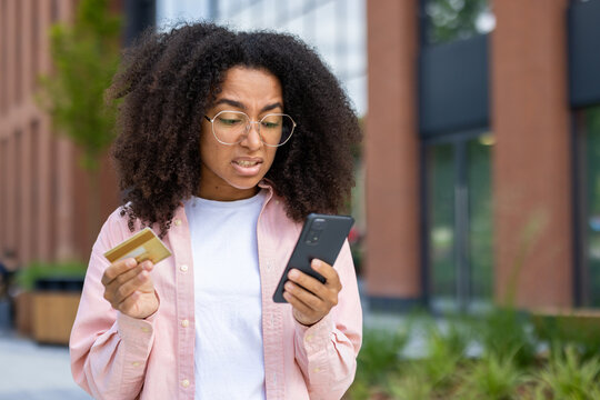 Anxious woman holding a credit card and smartphone outdoors, appearing concerned about online transactions or financial issues.