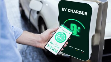 Businessman holding smartphone display battery status interface by smart EV mobile application while eco-friendly EV car recharging electricity from charging station in car park. Peruse