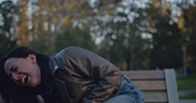 A young woman in a park displays a powerful emotional journey, transitioning from uncontrollable laughter and joy to profound sadness and crying while sitting on a bench.