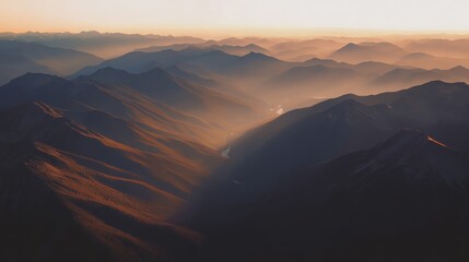Aerial view of mountain ranges illuminated by soft sunlight with misty valleys and a winding river creating a peaceful and scenic landscape at sunrise or sunset.