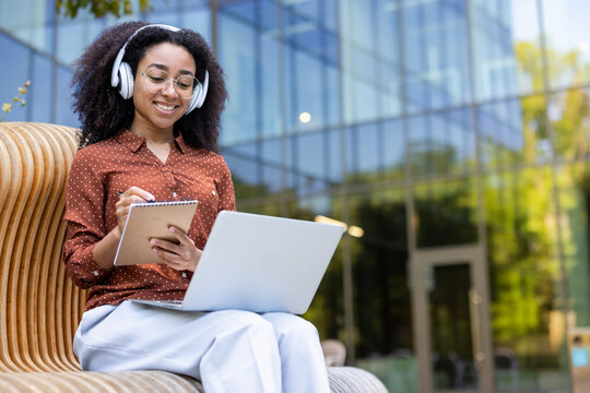 A smiling woman wearing headphones and glasses sits outside, taking notes on a notebook, and working on a laptop. - Powered by Adobe