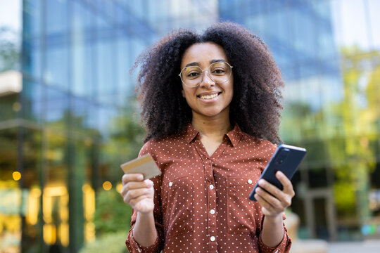 A smiling woman with curly hair holds a credit card and a smartphone outside a building, looking content. - Powered by Adobe