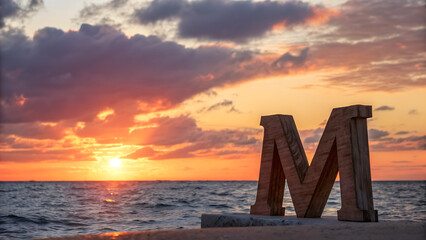 Letter m sculpture on a pier with a sunset over the ocean and cloudy sky in the background view - ai generated