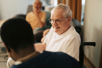 Smiling senior man sitting in wheelchair talking to male caregiver at retirement home