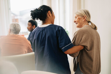 Rear view of female nursing assistant walking arm in arm with elderly woman at retirement home