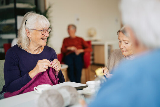 Senior female friends spending leisure time knitting while sitting at table in retirement home