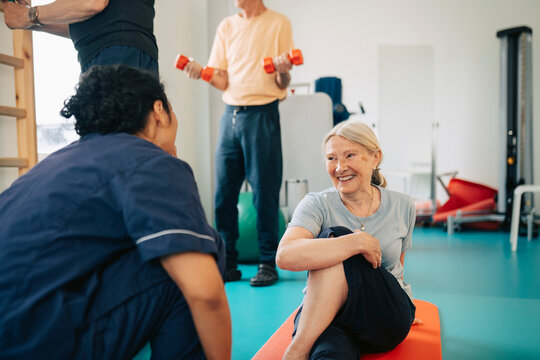 Smiling senior woman stretching and sitting on exercise mat while talking with therapist in rehabilitation center
