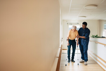 Male therapist assisting senior man walking with crutches in corridor at nursing home