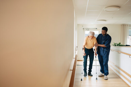 Male therapist assisting senior man walking with crutches in corridor at nursing home