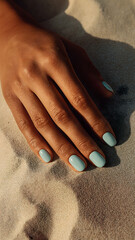 Close-up of a hand with light blue nail polish resting on warm beach sand, capturing a summery and relaxed coastal vibe.


