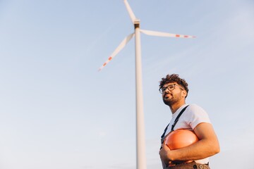 Young engineer holding helmet looking a wind turbine in the sky