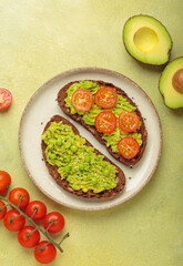 Two slices of whole rye bread are topped with mashed avocado, green peas, cherry tomatoes, and...