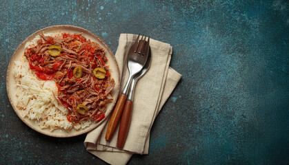 Ropa vieja, a traditional Cuban dish, is served with white rice, tomato sauce, and green olives on a plate from above on dark rustic background, copy space