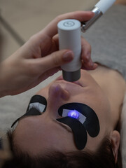 A master extends a woman's eyelashes using an LED lamp. Vertical photo. 