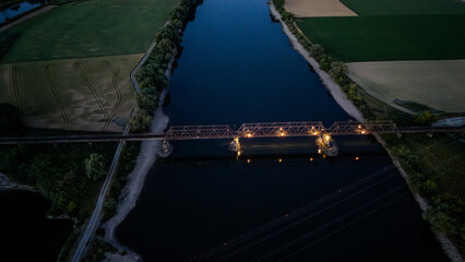 Blick auf eine beleuchtete Eisenbahnbrücke über einen ruhigen Fluss bei Dämmerung